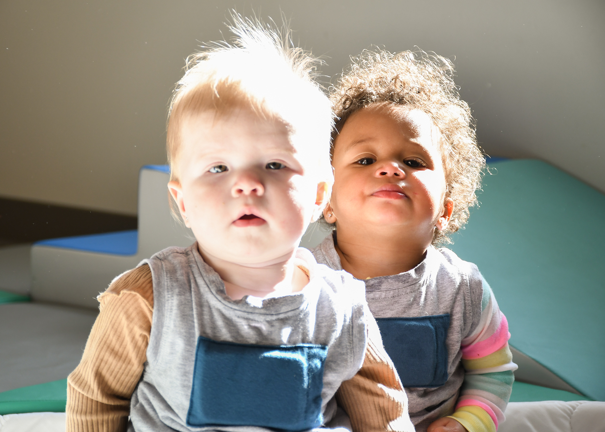 A blonde white baby and a brown, African American baby with curly hair sit on the floor in a sunlight room.
