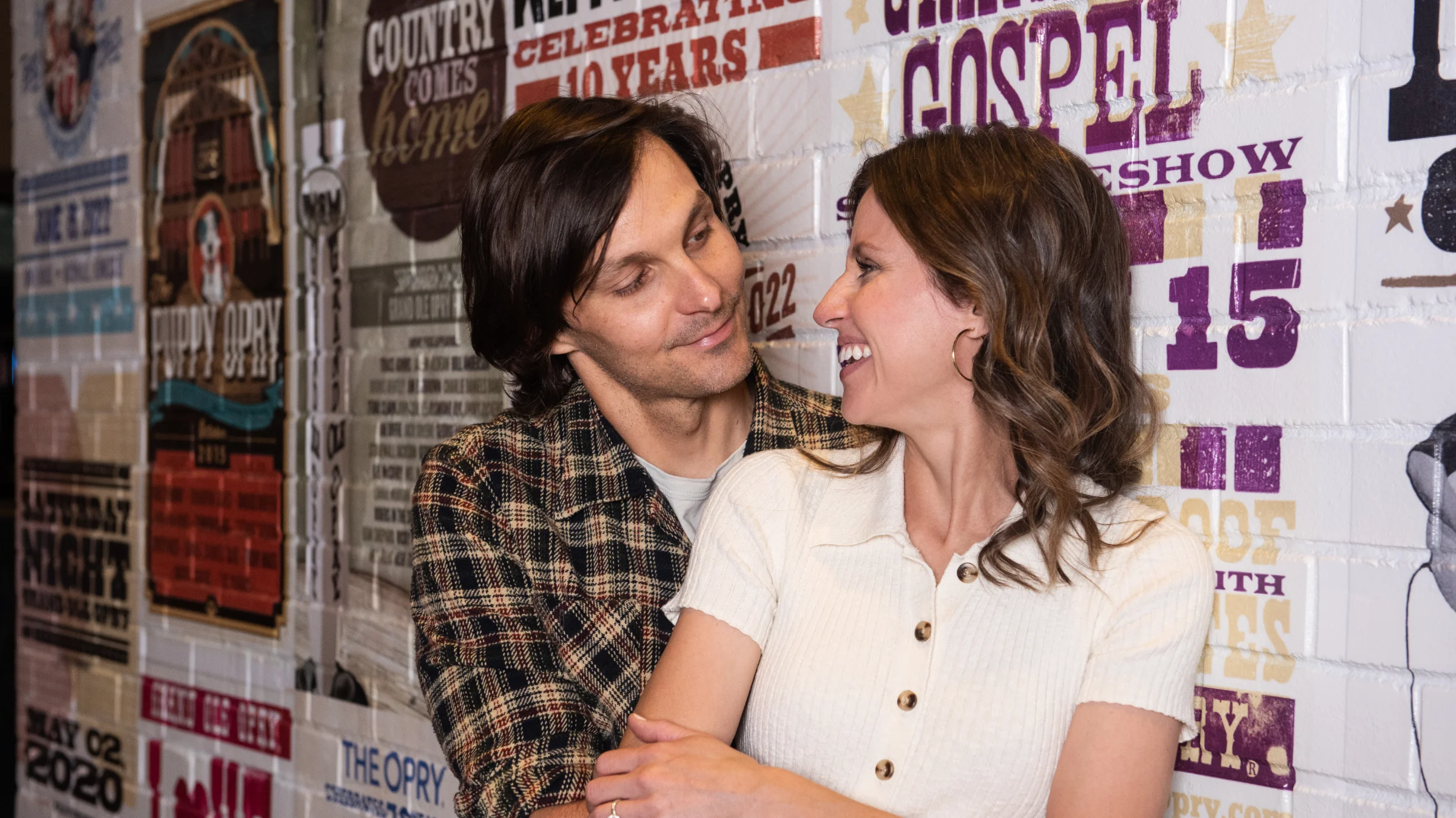 Kristen and Charlie Worsham embrace and smile at each other in front of a wall of painted posters.
