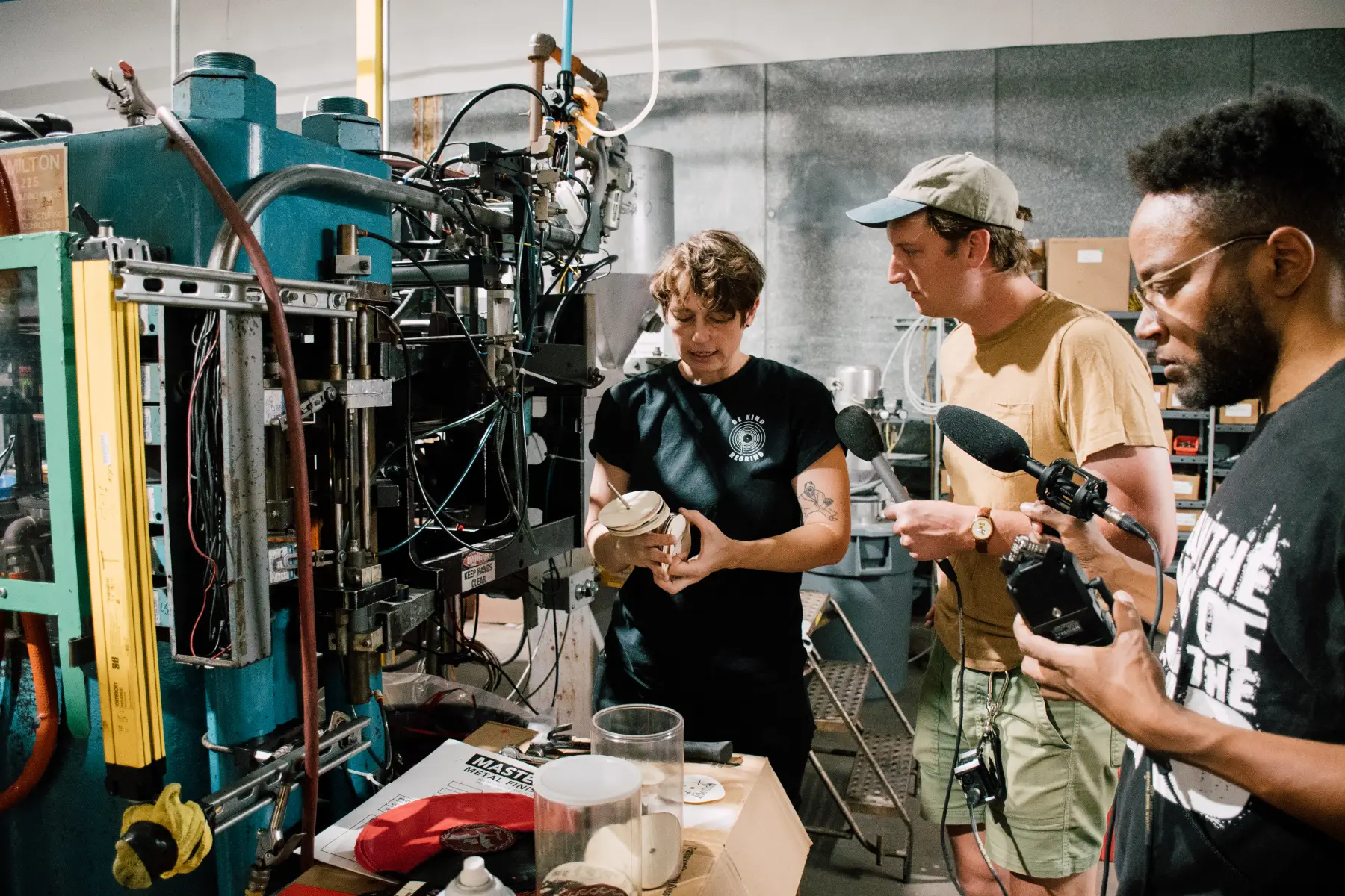 An artist explains a commercial pressing machine to a reporter and sound technician