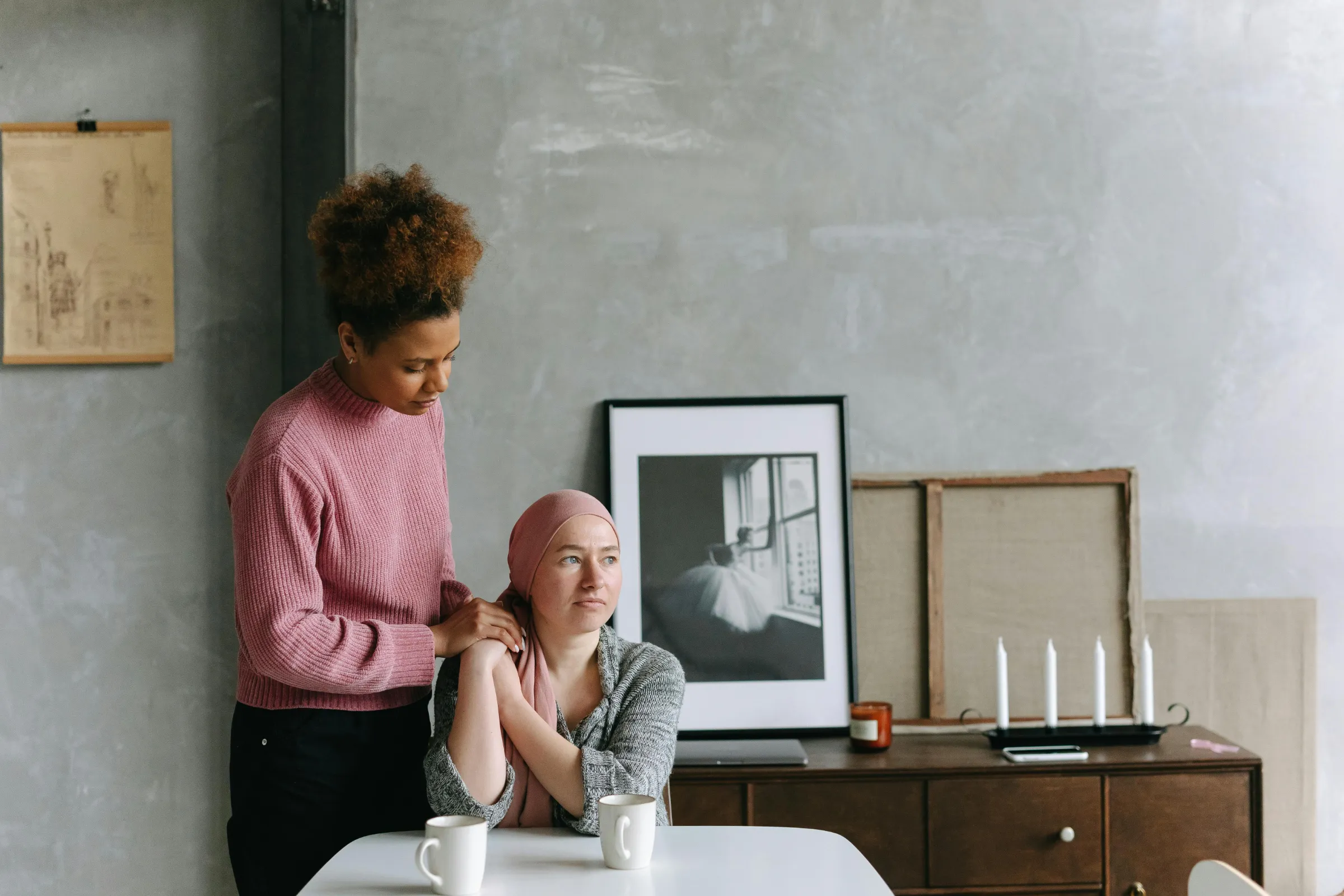 A young woman stands comforting another who sits at a table staring off into the distance.