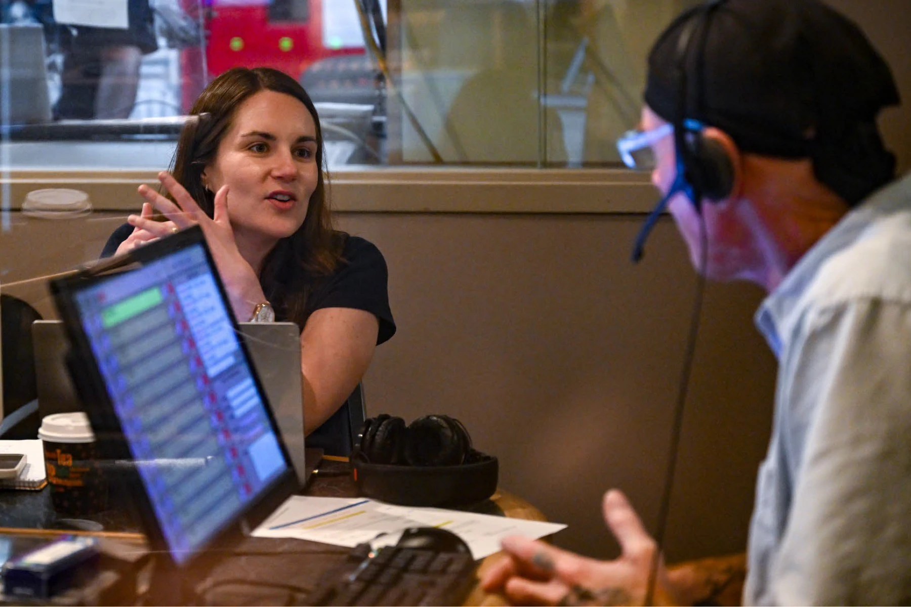 A talk show host wearing a headset in front of a audio console interviews another individual inside a studio.