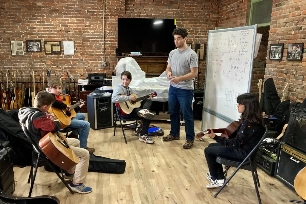 A student plays guitar during a class at The Quest Center surrounded by other students, the teacher, and various instruments.