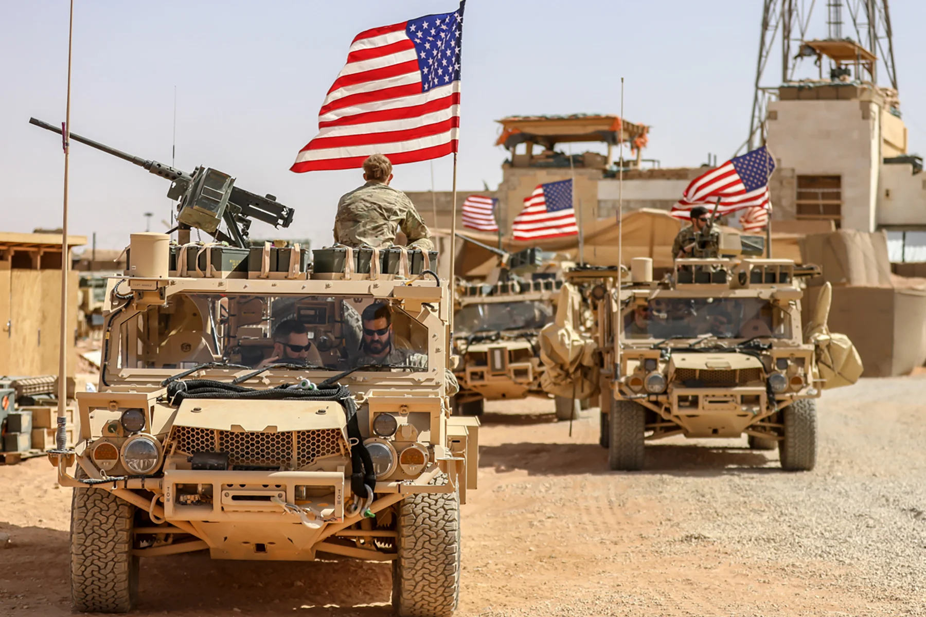 A line of 3 US armored humvee trucks with soldiers on top and US flags waving as they sit outside an outpost in the Middle East