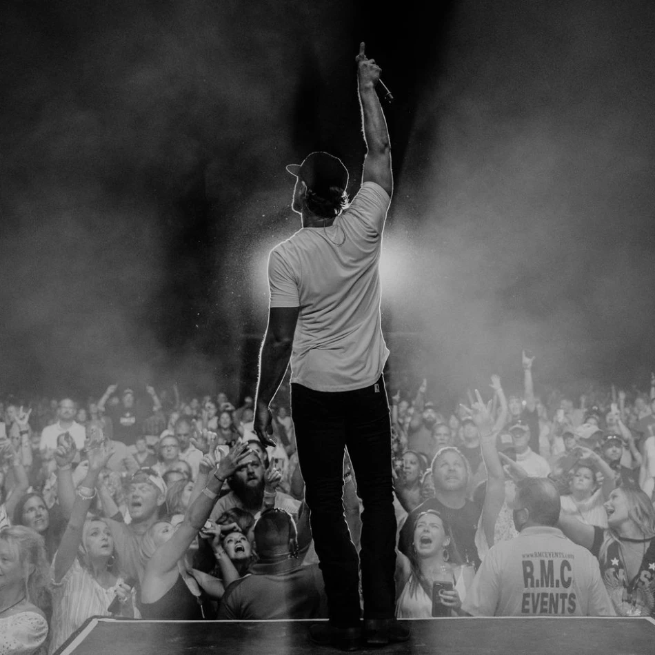A black and white image of an artist standing in front of a crowd raising his hand and pointing to the sky with a backlight behind him.