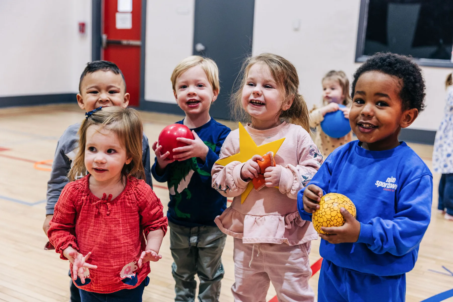 A group of children smiling holding balls and toys inside a gym during playtime.