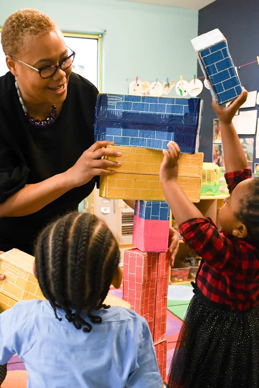 A teacher helps two small children stack cardboard blocks painted to looked like colored bricks inside a classroom.