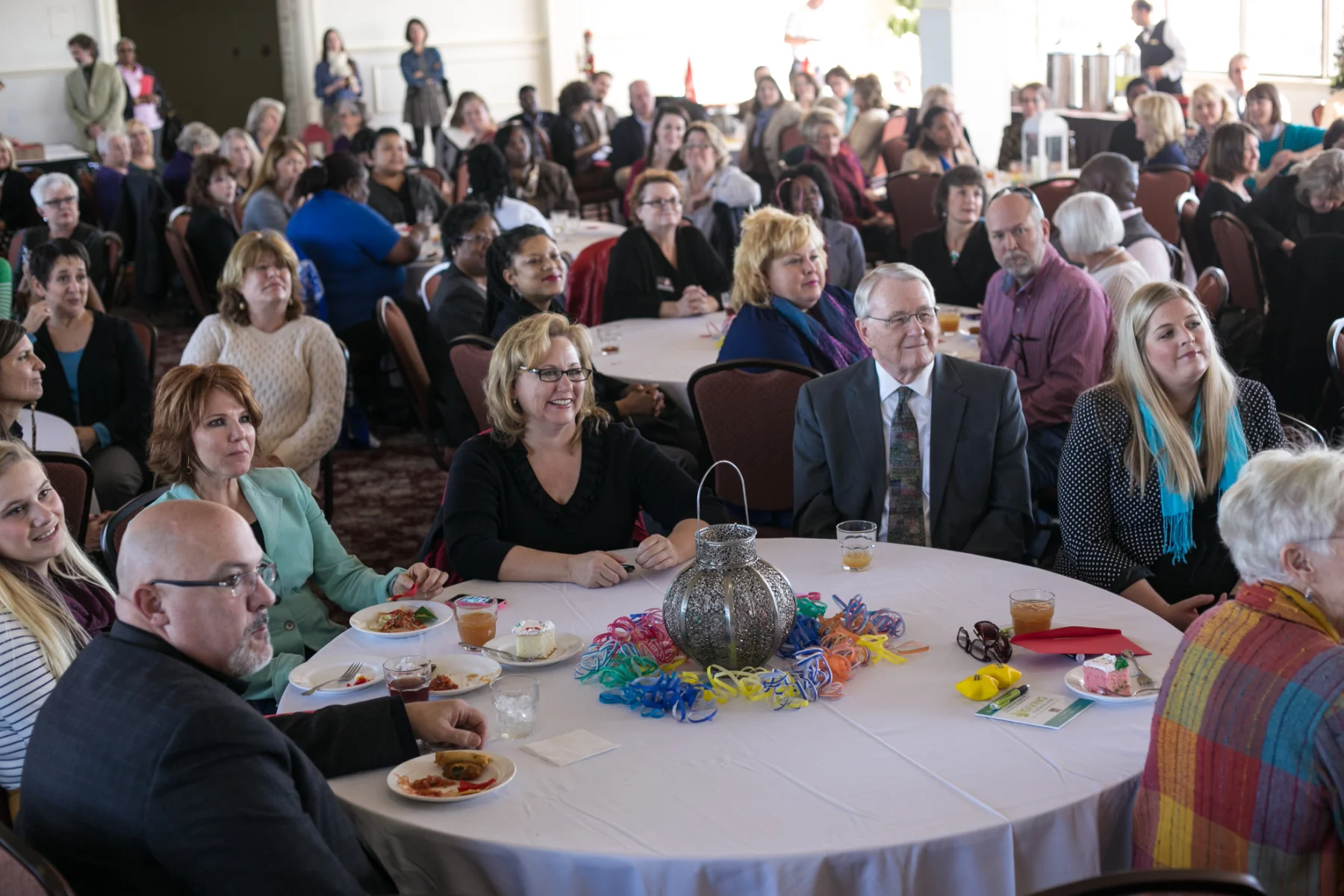 A ballroom at a CFMT event filled with tables, food & drinks, and guests looking off screen at a speaker.