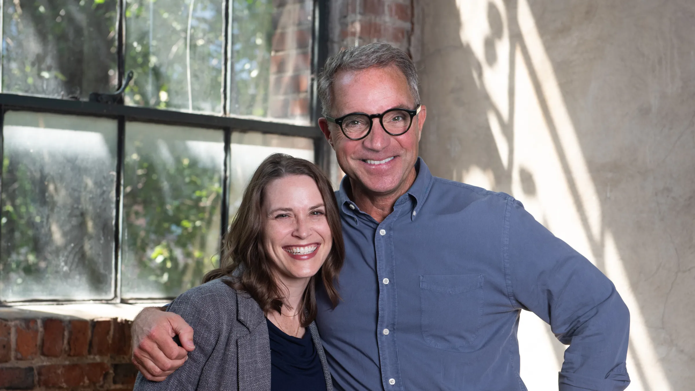Hal Cato and Stephanie Coleman share a hug inside an industrial style office with a brick framed window behind them.